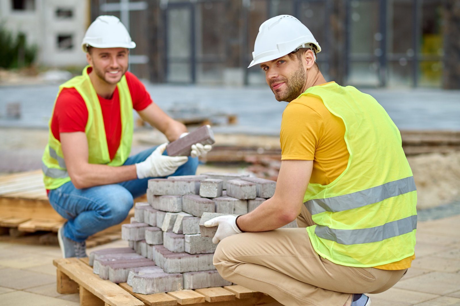Two workers stacking bricks looking at camera Manual labour. Two young adult smiling workers in protective helmet and gloves crouching stacking bricks looking at camera at construction site on warm day